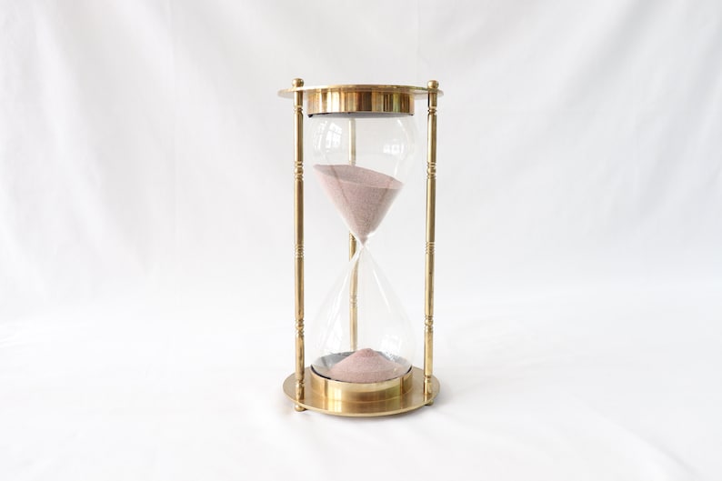 May include: A brass hourglass with a clear glass bulb and pink sand. The hourglass is on a white surface.