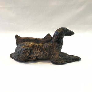 May include: A bronze-colored cast iron sculpture of a dog lying down. The dog has a long, slender body and a long, pointed snout. The sculpture is detailed with a textured finish.