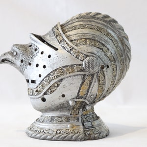 May include: A silver helmet with an eagle head design and intricate details. The helmet is decorated with gold accents and has a textured surface.