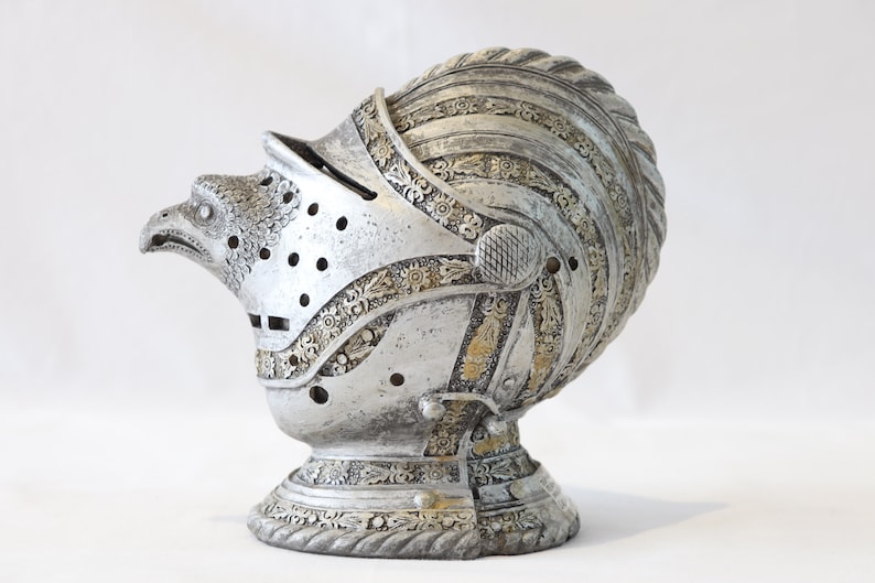 May include: A silver helmet with an eagle head design and intricate details. The helmet is decorated with gold accents and has a textured surface.