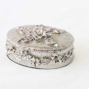 May include: A silver trinket box with a floral design and pink rhinestones. The box is oval shaped and has a raised floral design on the lid.