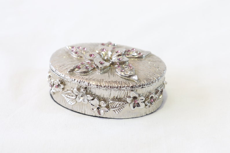 May include: A silver trinket box with a floral design and pink rhinestones. The box is oval shaped and has a raised floral design on the lid.