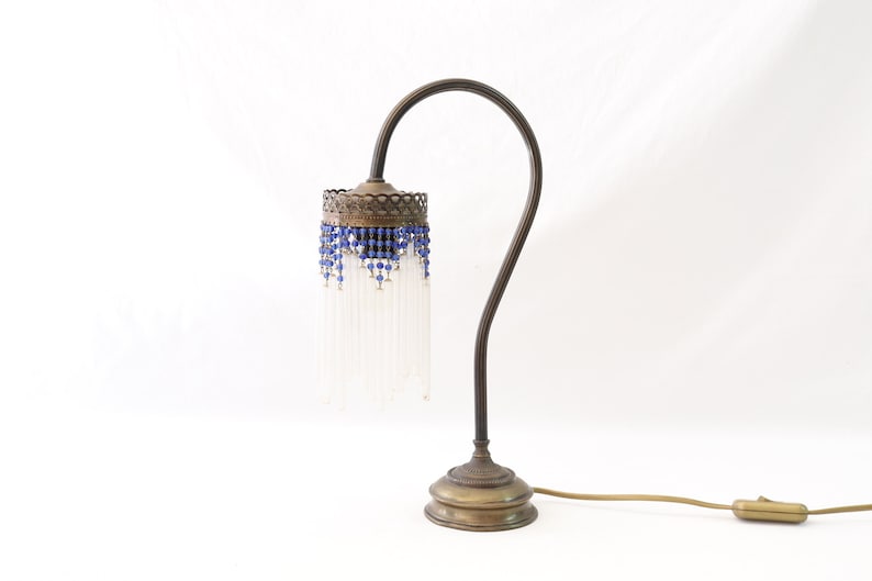 K&ouml;nnte beinhalten: Eine Vintage-Tischlampe aus Messing mit einem wei&szlig;en Perlenlampenschirm und blauen Akzenten. Die Lampe hat einen flexiblen Arm und einen runden Sockel.