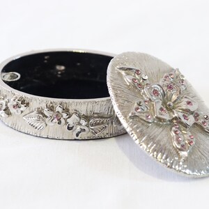 May include: Silver trinket box with a floral design. The lid is decorated with a large flower with pink rhinestones. The box is open and the inside is black.