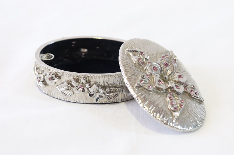 May include: Silver trinket box with a floral design. The lid is decorated with a large flower with pink rhinestones. The box is open and the inside is black.