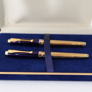 May include: A set of two pens in a blue velvet box. The pens have gold trim and a purple barrel.