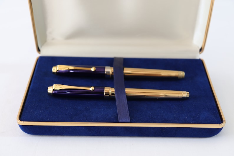 May include: A set of two pens in a blue velvet box. The pens have gold trim and a purple barrel.