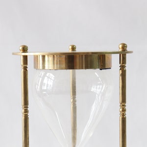 May include: A brass hourglass with a clear glass bulb. The hourglass is standing upright on a white surface.