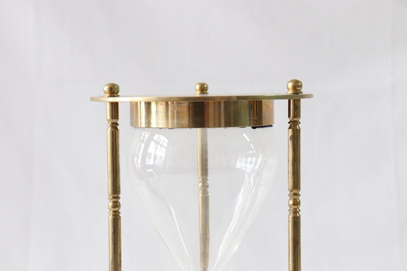 May include: A brass hourglass with a clear glass bulb. The hourglass is standing upright on a white surface.