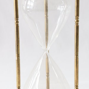 May include: A brass hourglass with a clear glass bulb. The hourglass is standing upright on a white surface.