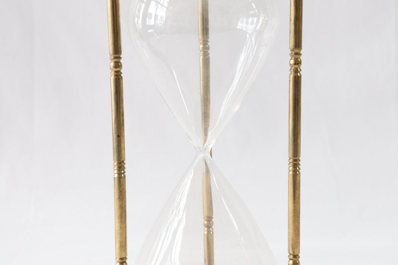 May include: A brass hourglass with a clear glass bulb. The hourglass is standing upright on a white surface.