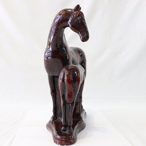 May include: A dark brown ceramic sculpture of a horse and foal standing side by side. The horse is standing with its head turned to the side, while the foal is standing with its head down. The sculpture is on a white background.