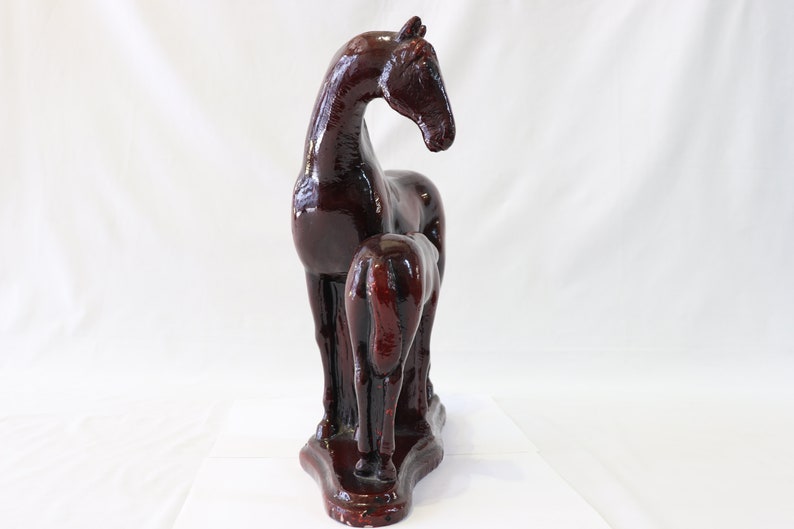 May include: A dark brown ceramic sculpture of a horse and foal standing side by side. The horse is standing with its head turned to the side, while the foal is standing with its head down. The sculpture is on a white background.