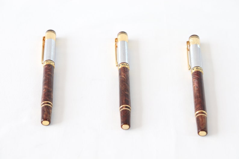 May include: Three brown and silver pens with gold accents. The pens have a wood grain pattern and are arranged in a row on a white surface.