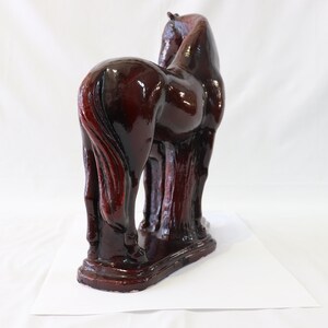 May include: A dark brown ceramic horse figurine with a glossy finish. The horse is standing on all four legs with its head turned to the side. The figurine is detailed with a flowing mane and tail.
