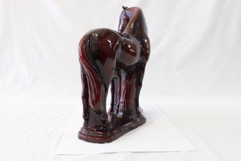 May include: A dark brown ceramic horse figurine with a glossy finish. The horse is standing on all four legs with its head turned to the side. The figurine is detailed with a flowing mane and tail.