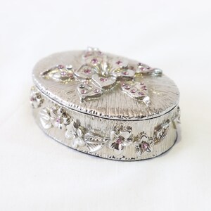 May include: A silver oval trinket box with a floral design and pink rhinestones. The lid features a butterfly with pink rhinestones.