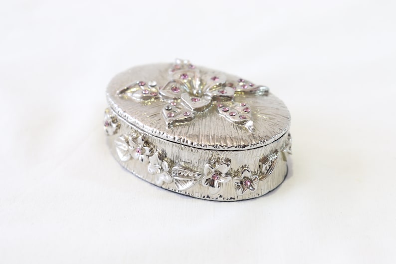 May include: A silver oval trinket box with a floral design and pink rhinestones. The lid features a butterfly with pink rhinestones.