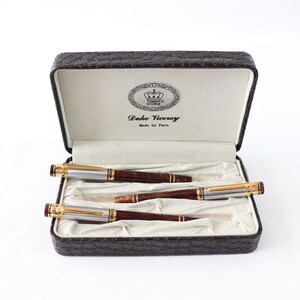 May include: A set of three pens in a brown leather case. The pens are silver with gold accents and have a wood grain design. The case has a gold emblem with the words "Duke Viceroy" and "Made by Paro".