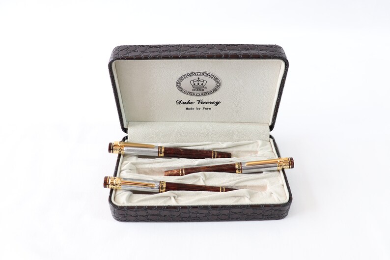 May include: A set of three pens in a brown leather case. The pens are silver with gold accents and have a wood grain design. The case has a gold emblem with the words "Duke Viceroy" and "Made by Paro".