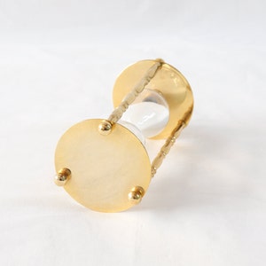 Pu&ograve; includere: Una clessidra color oro con un centro in vetro trasparente riempito di sabbia bianca. La clessidra ha una base circolare con tre piedini e una struttura di supporto decorativa e attorcigliata.