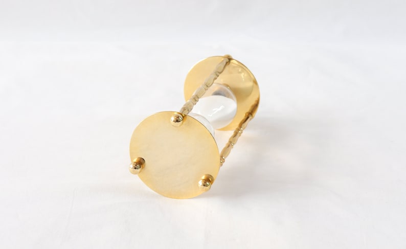Pu&ograve; includere: Una clessidra color oro con un centro in vetro trasparente riempito di sabbia bianca. La clessidra ha una base circolare con tre piedini e una struttura di supporto decorativa e attorcigliata.
