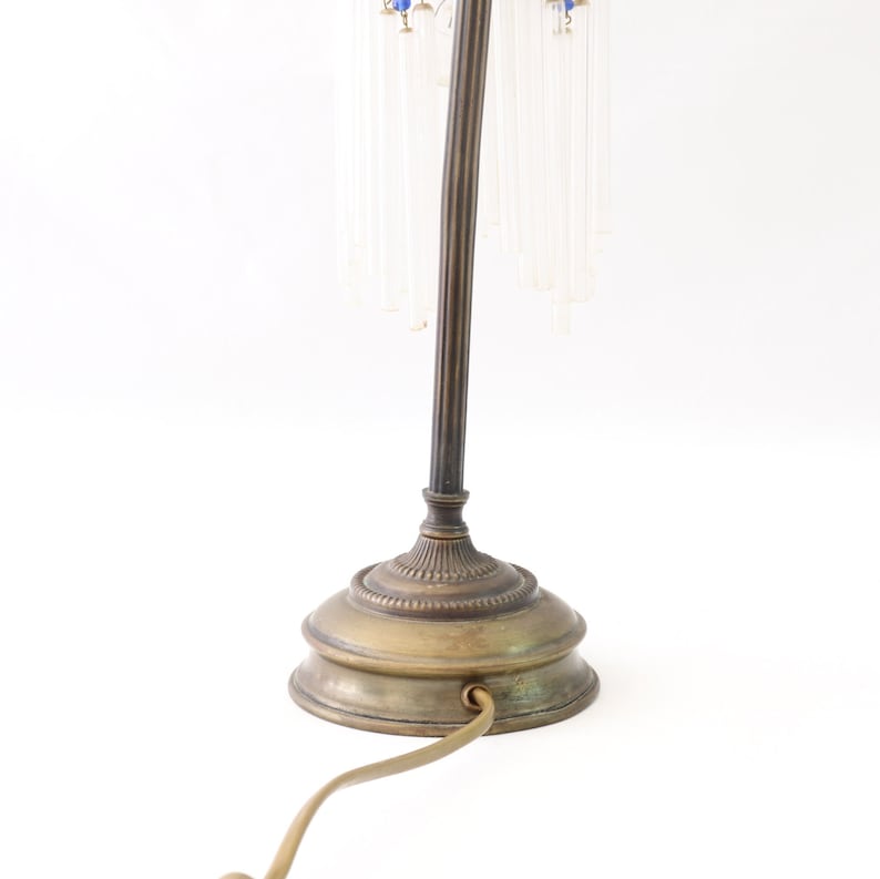 K&ouml;nnte beinhalten: Eine Tischlampe aus Messing mit einem gebogenen Schaft und einem runden Sockel. Die Lampe hat einen blauen und klaren Glasschirm mit kristallartigen Str&auml;ngen, die von unten h&auml;ngen.