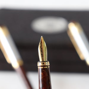 May include: Close-up of a gold fountain pen nib with a brown and gold marbled pen barrel. The pen is in focus, while two other pens are blurred in the background.