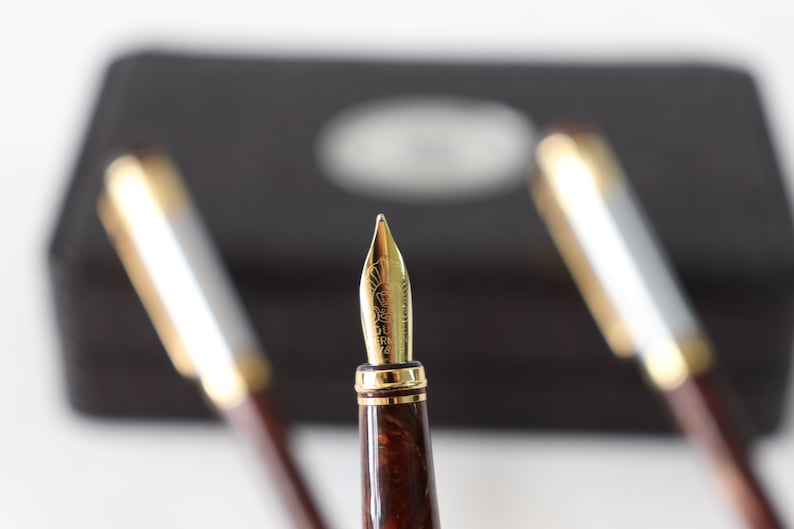 May include: Close-up of a gold fountain pen nib with a brown and gold marbled pen barrel. The pen is in focus, while two other pens are blurred in the background.