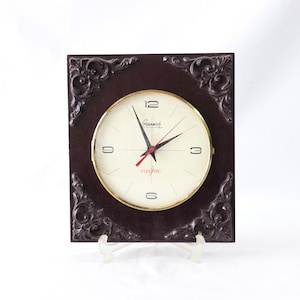 May include: Square brown wall clock with a cream-colored face and gold trim. The clock has black hands and a red second hand. The clock face has black numbers and the word "ELECTRIC" in red. The clock is supported by a clear stand.