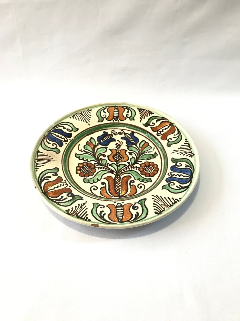 May include: A white ceramic plate with a green, blue, and orange floral design. The plate has a white background with a green border and a floral design in the center.
