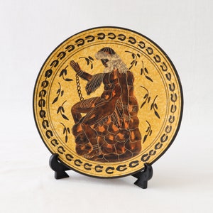 May include: A decorative plate featuring a classical figure in brown and orange tones, chained and seated on a rock formation. The plate has a yellow background with a black border and leaf design. It is displayed on a black stand.