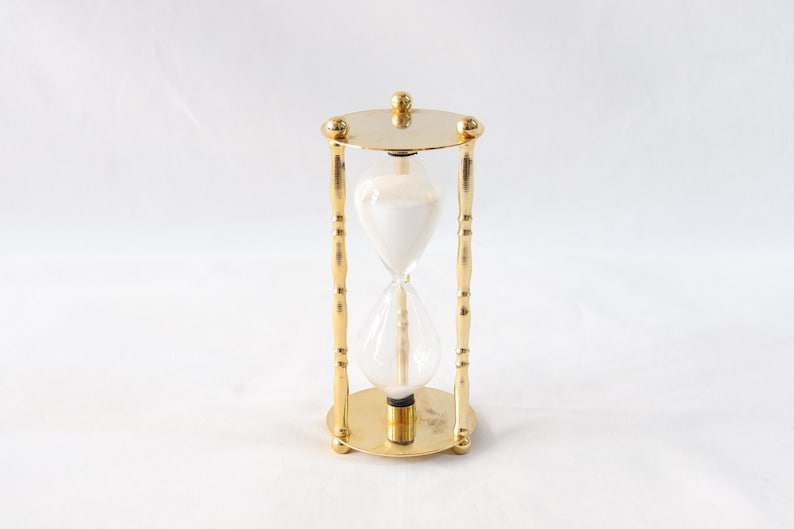 Pu&ograve; includere: Una clessidra color oro con un centro in vetro trasparente riempito di sabbia bianca. La clessidra &egrave; sostenuta da una struttura in metallo color oro con base e parte superiore rotonde.