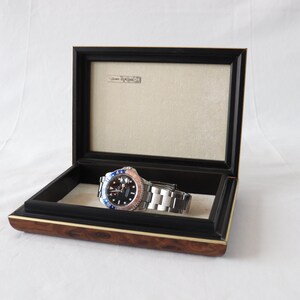 May include: A silver watch with a black face and a blue and red bezel is displayed in a wooden box with a black velvet lining. The box has a gold trim.