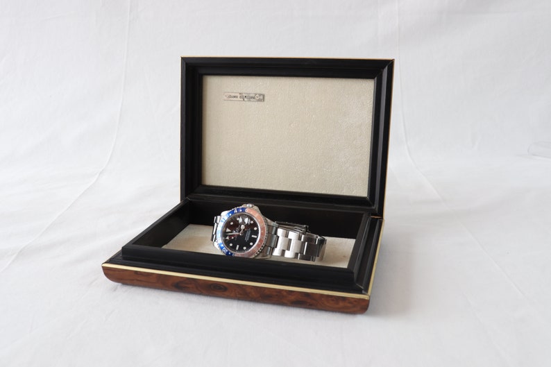 May include: A silver watch with a black face and a blue and red bezel is displayed in a wooden box with a black velvet lining. The box has a gold trim.