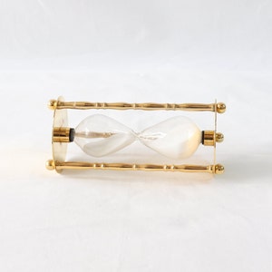 Pu&ograve; includere: Una clessidra con un centro in vetro trasparente e una cornice in metallo color oro. La clessidra ha un design classico con una parte superiore e inferiore bulbosa, collegate da un centro stretto. La cornice presenta barre decorative in stile bamb&ugrave; e terminali circolari.