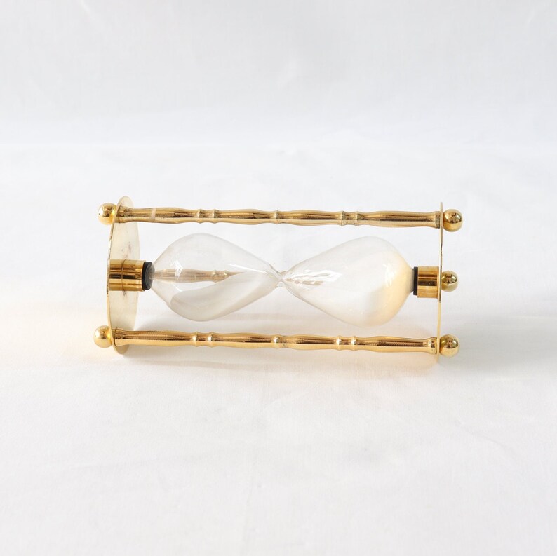 Pu&ograve; includere: Una clessidra con un centro in vetro trasparente e una cornice in metallo color oro. La clessidra ha un design classico con una parte superiore e inferiore bulbosa, collegate da un centro stretto. La cornice presenta barre decorative in stile bamb&ugrave; e terminali circolari.
