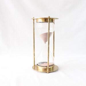 May include: A brass hourglass with a clear glass bulb and pink sand. The hourglass is standing on a white surface.