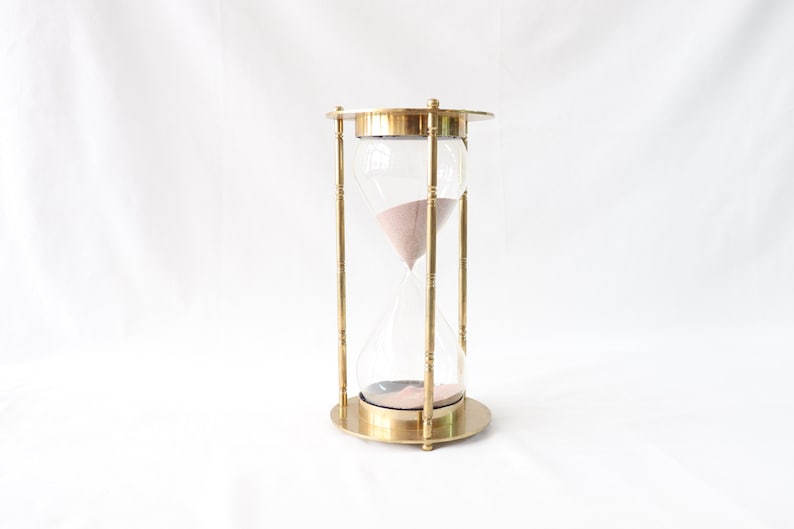 May include: A brass hourglass with a clear glass bulb and pink sand. The hourglass is standing on a white surface.