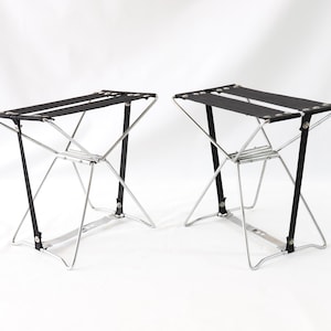 Könnte beinhalten: Zwei schwarze Klapphocker mit silbernen Metallrahmen. Die Hocker sind zusammengeklappt und stehen auf einer weißen Oberfläche.