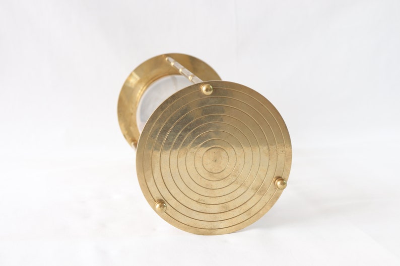 May include: Brass hourglass with a circular base and a clear glass hourglass. The base has a concentric circle design.