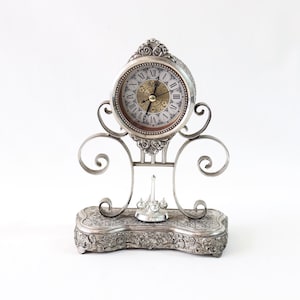 Può includere: Un'antica pendola argentata con un quadrante rotondo con numeri romani e dettagli ornati. L'orologio ha una base decorativa e un design delicato e a volute, che mostra un'estetica vintage.