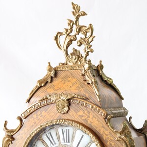 May include: A decorative clock with a gold-toned finish and intricate details. The clock face is surrounded by a wooden frame with a marquetry design. The clock has Roman numerals and a gold-toned hand.