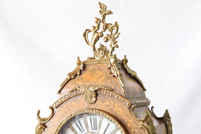 May include: A decorative clock with a gold-toned finish and intricate details. The clock face is surrounded by a wooden frame with a marquetry design. The clock has Roman numerals and a gold-toned hand.