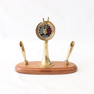May include: A gold-toned desk organizer with a ship's telegraph design and two pen holders. The telegraph features a black and white dial with red markings and the text "Engine Room Telegraph".