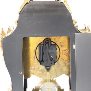 May include: Back view of a gold-toned clock with a black face and ornate details. The clock has a quartz movement and is made by the brand "World Quartz".