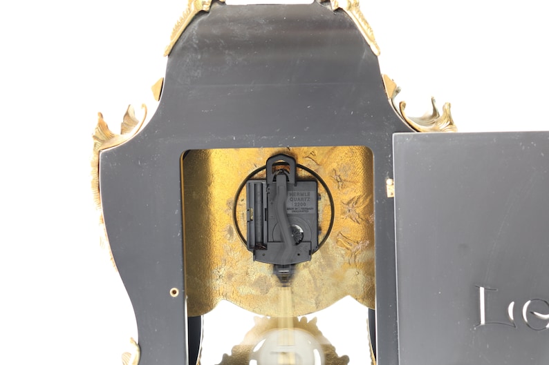 May include: Back view of a gold-toned clock with a black face and ornate details. The clock has a quartz movement and is made by the brand "World Quartz".