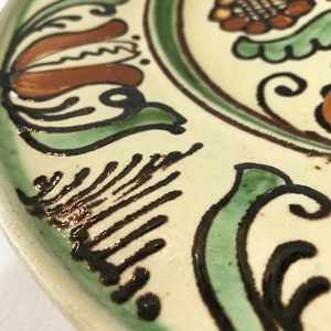 May include: A white ceramic plate with a green, brown, and black floral design. The plate has a raised rim and a glossy finish.