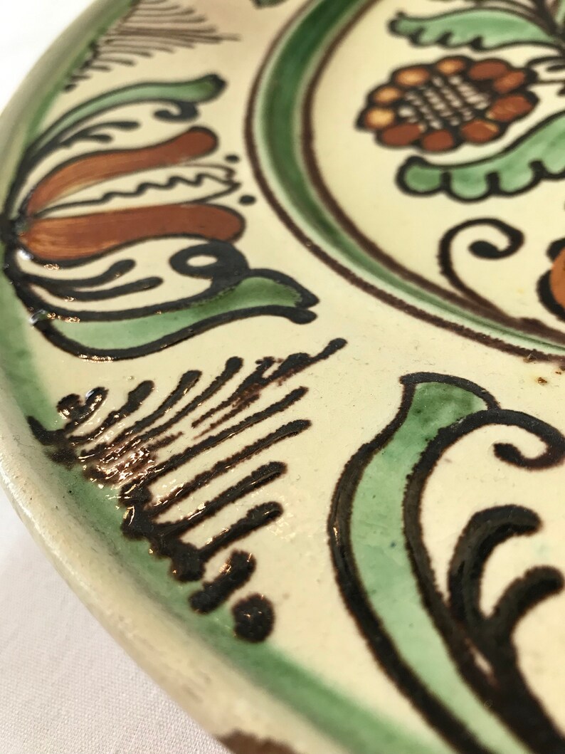 May include: A white ceramic plate with a green, brown, and black floral design. The plate has a raised rim and a glossy finish.
