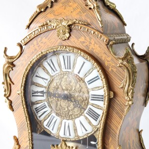 May include: A gold and brown ornate clock with Roman numeral markings on the face. The clock has a decorative design with intricate details and a glass cover over the face.